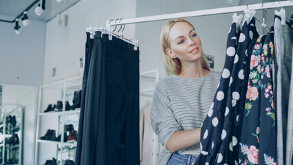 Pretty blond girl is choosing fashionable clothes in boutique. She is looking through fine garments...