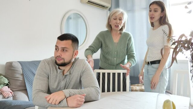 Portrait of displeased young adult man sitting at home ignoring his wife and mother-in -law berating him