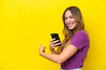 Young caucasian woman isolated on yellow background using mobile phone and pointing back