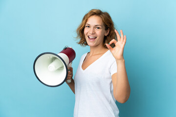 Young Georgian woman isolated on blue background holding a megaphone and showing ok sign with...