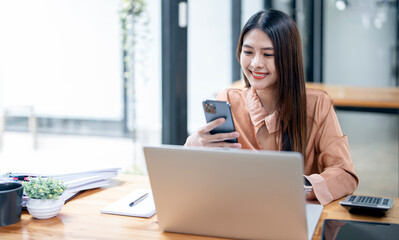 Happy young asian businesswoman using mobile phone, standing at desk with laptop computer.