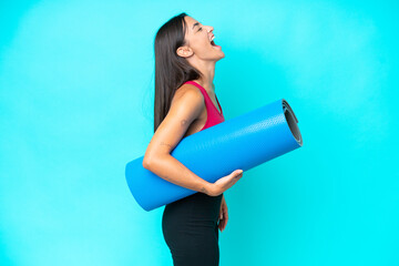 Young sport caucasian woman going to yoga classes while holding a mat isolated on blue background...