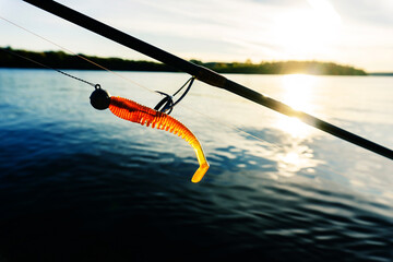 A fragment of a spinning rod and a silicone bait against the background of the river and the sun's...