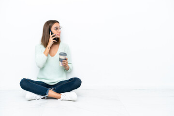 Young caucasian woman sitting on the floor isolated on white background holding coffee to take away and a mobile