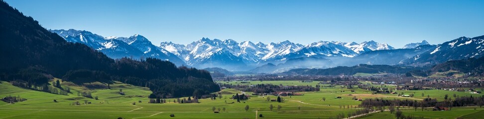 Panoramic view of a green meadow surrounded by snow-covered mountains  on a sunny day
