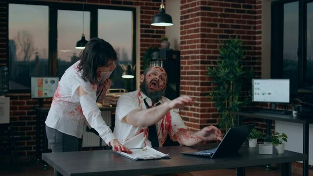 Creepy mindless zombie sitting in office while working on modern laptop. Spooky brain dead monster with deep and bloody scars typing on portable computer while dead walking coworker brings clipboard.