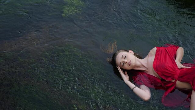 Art beautiful romantic portrait of a sexy young woman in a red dress, lying in a river with green algae in summer in nature during the day. photo shoot of a beautiful young model