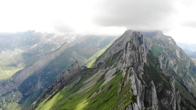 Aerial flyover over and away from the cliffs of Schafler ridge in Appenzell, Switzerland with cliffs, mountain peaks and lush summer green mountainside in view