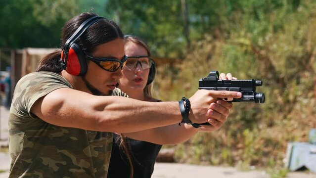 White man in camo t-shirt safety headphones and goggles holding handgun with female instructor showing how to aim it. Firearm training ar firing range. Outdoor horizontal shot. High quality 4k footage