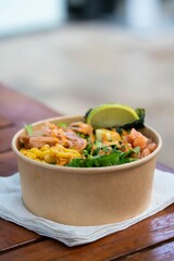 Vertical shot of a paper bowl of delicious poke with corn, salmon, seaweed, lime, and spicy mayo