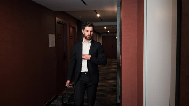 A Man In A Classic Suit With A Suitcase Walks Down The Corridor In The Hotel, Approaches The Door And Takes Out The Electronic Key For Opening Door. Businessman Arriving In Hotel For Business Trip.