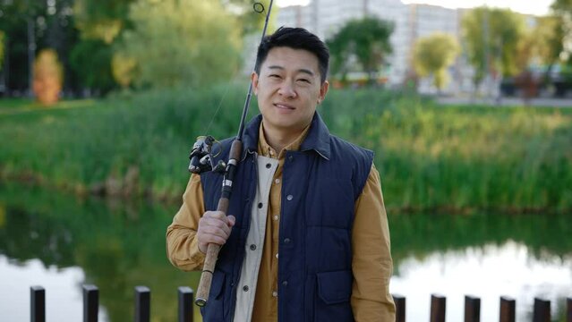 Smiled asian fisherman wearing waistcoat holding the fishing rod standing looking at the camera on the blurred local lake background. People and leisure time, fishing concept