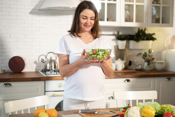 Beautiful mama with salad standing in bright light kitchen inside cozy flat.