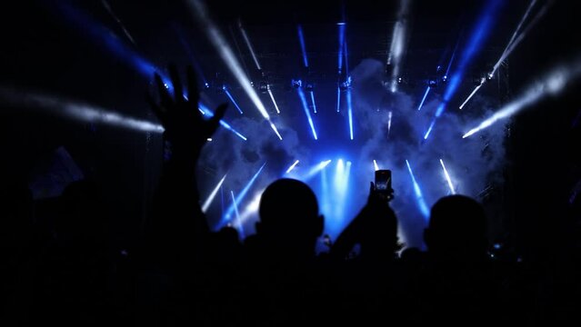 Silhouettes of exited fans in shiny concert stage with DJ performing on a night party