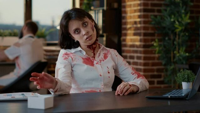 Weird looking spooky zombie woman waving at camera while sitting in office. Mindless brain-eating undead monster with bloody wounds sitting at table in workspace while waving hand.