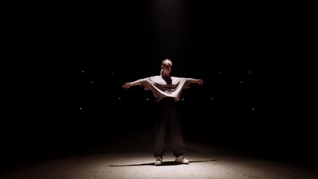 brash hip-hop dancer girl is moving in soffit light in dark studio or stage, street dance battle