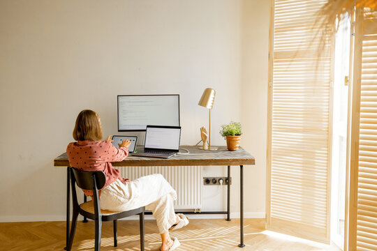 Woman works on computers while sitting by a cozy workplace in sunny living room at home. View from the backside. Concept of remote work from home office. Mockup image