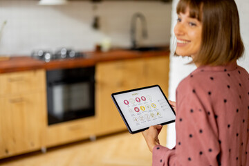 Woman holds a digital tablet with running mobile application for smart home, standing in kitchen of modern apartment. Controling smart devices with a digital tablet