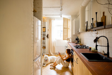 Woman uses smart phone while sitting with her dog in kitchen. Interior view on modern and stylish kitchen in beige tones. Domestic lifestyle concept