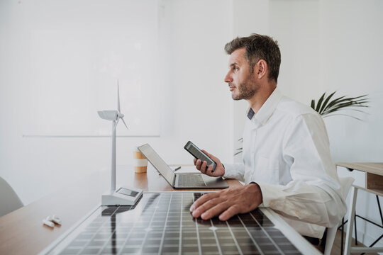 Engineer with hand on solar panel by wind turbine model at desk