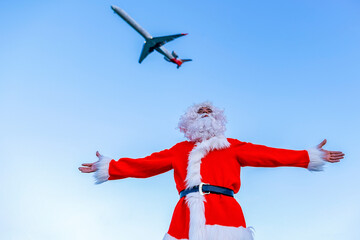 Man wearing Santa Claus costume standing with arms outstretched below flying airplane