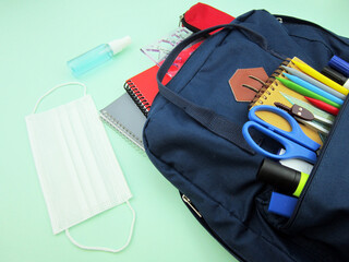    As COVID-19 prevention goes back to school and new normal concept. Top view of backpack with school supplies, mask and disinfectant gel on green background.                           