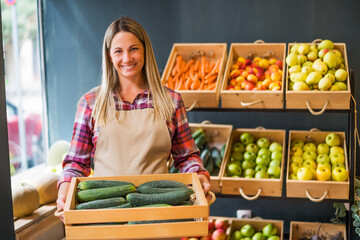 Woman works in fruits and vegetables shop. She is holding basket with cucumber.