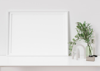 Empty horizontal picture frame standing on white shelf. Frame mock up. Copy space for picture, poster. Template for your artwork. Close up view. Plant in vase, home accessories, sculpture. 3D render.
