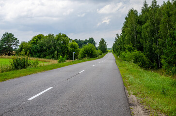 View of the paved road which runs through the fields and forest in the daytime.