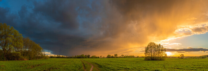 Stunning panorama of a dramatic sunset over a summertime field and forest, with heavy storm clouds in the background.