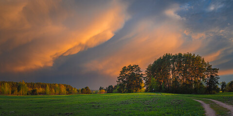 Wide angle view of dramatic sunset sky over summer field and forest with orange and blue storm clouds
