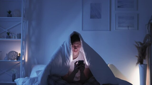 Mobile fatigue. Gadget insomnia. Social media addiction. Exhausted bored sleepy yawning man using phone chatting late under blanket on bed in dark night bedroom.