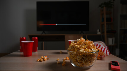 Glass bowl of popcorn and cups and remote control on wooden table. Family vacation, recreation, relaxation and hobby concept