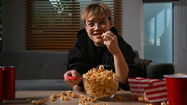 Happy man with remote control eating popcorn and watching movie at night. Entertainment and leisure activity concept