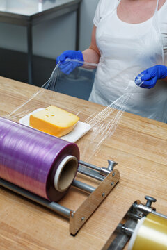 Female worker wraps piece of cheese with transparent film that she pulls from large roll.