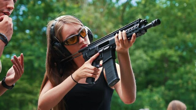 caucasian woman wearing safety gear aiming submachine gun observed by male instructor in camo t-shirt. Firearms training at shooting range. Outdoor horizontal shot. High quality 4k footage