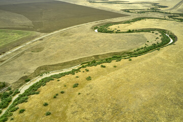a winding muddy river with overgrown green banks in a sun-scorched steppe rural landscape