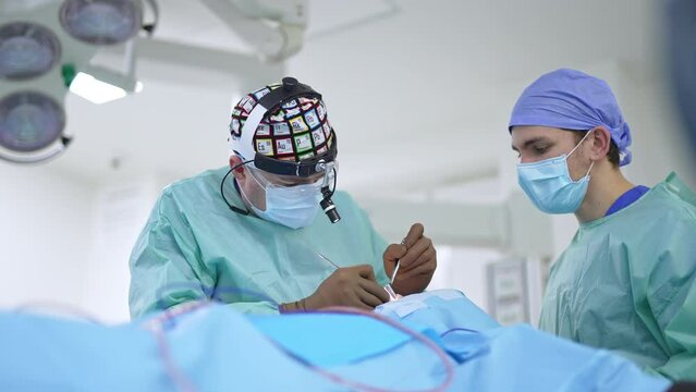 Male surgeon in funny cap and device glasses working hard at operation. Male assistant helping the doctor.