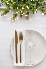 Festive table setting with gold cutlery , porcelain plate, wine glass and centerplace on white tablecloth with gold bokeh.