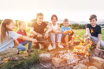Six kids group Boys and girls cheerfully laughed and roasted sausages on sticks over a campfire flame near the green tent. Outdoor active time spending or camping in Nature concept. - obrazy, fototapety, plakaty