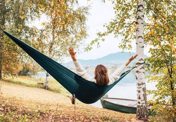 The young woman cheerfully rose arms up while she swinging in a hammock between the birch trees on the mountain lake bank. Out-of-town Outdoor Recreation in Nature concept image.
