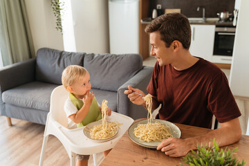 Upper view of handsome dad and cute blond baby in high chair eating spaghetti for lunch at kitchen...