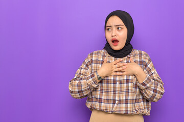 Fototapeta premium Amazed young Asian woman in plaid shirt hands on chest, looking away at copy space with wow face expression isolated on purple background
