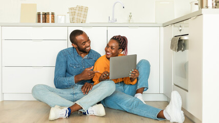 Happy black wife and husband using laptop, watch video or shopping online in kitchen interior, sitting together on floor