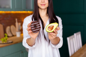a young woman in a white shirt stands in the kitchen with a donut in one hand and an avocado in the...