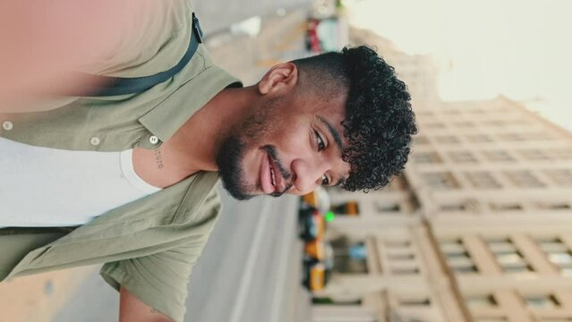VERTICAL VIDEO: Young happy man with beard dressed in an olive color shirt stands next to the road uses smartphone, smiles and makes video call