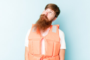 Young caucasian man wearing life jacket isolated on blue background dreaming of achieving goals and...