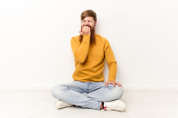 Young caucasian man sitting on the floor isolated on white background biting fingernails, nervous and very anxious.