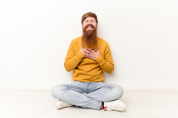 Young caucasian man sitting on the floor isolated on white background laughs out loudly keeping hand on chest.