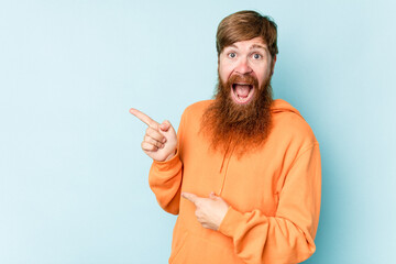 Young caucasian red-haired man isolated on blue background excited pointing with forefingers away.